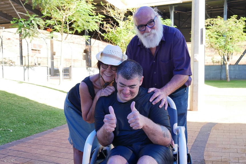 A man sits with two thumbs up as an older woman and man hug him from behind and smile broadly