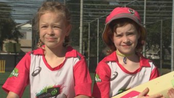 Two young girls dressed up in bright pink cricket gear, holding a bat