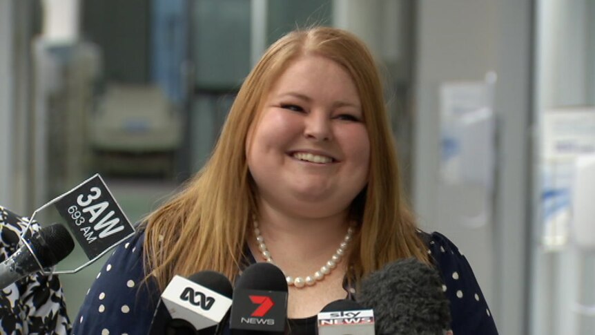 A smiling woman with red hair addresses the media in a hospital corridor.