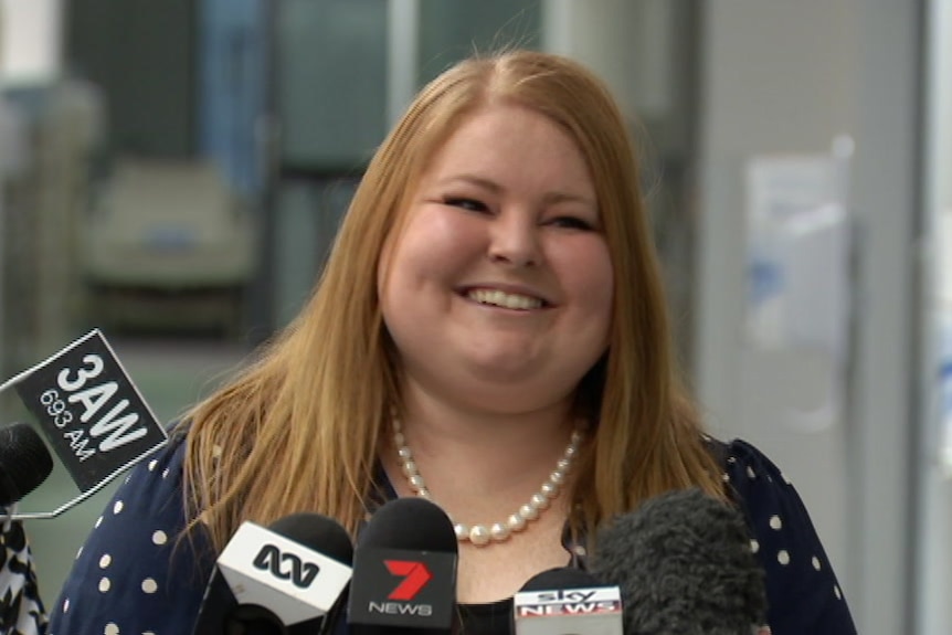 A smiling woman with red hair addresses the media in a hospital corridor.