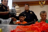 Two accused drug smugglers, sit in orange prison jumpsuits in front of drugs, with several police officers behind them