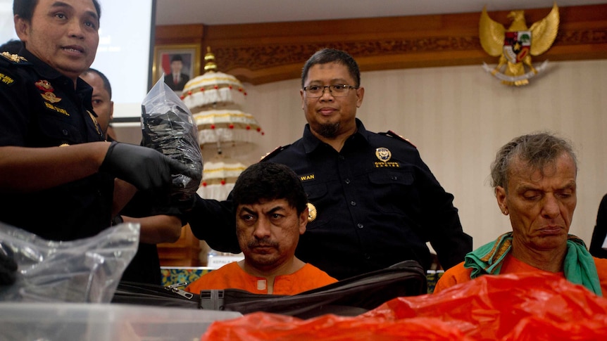 Two accused drug smugglers, sit in orange prison jumpsuits in front of drugs, with several police officers behind them