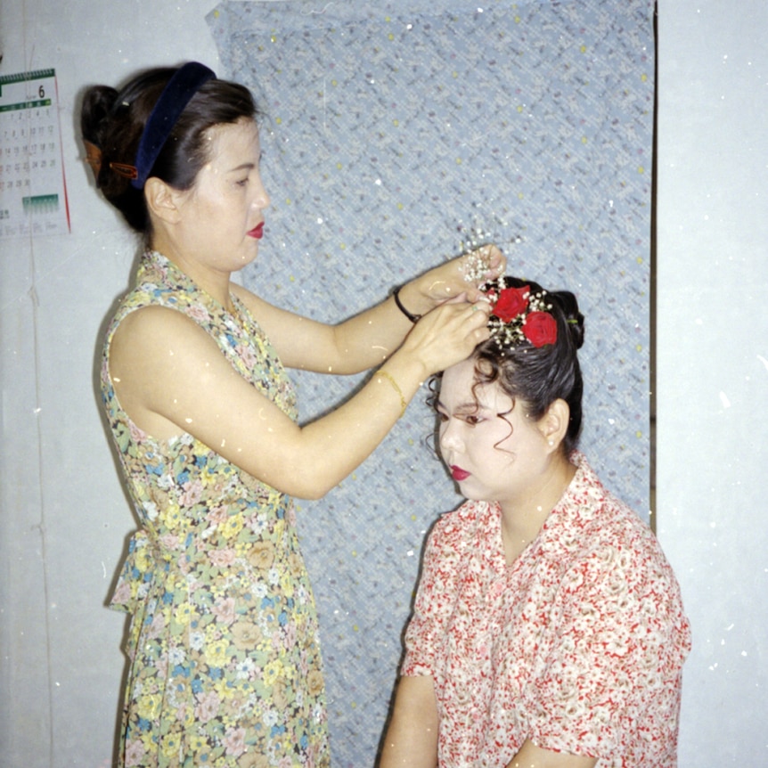 You view a woman in a floral dress applying a hairpiece to a woman sitting down, with a face full of white makeup and red lips.