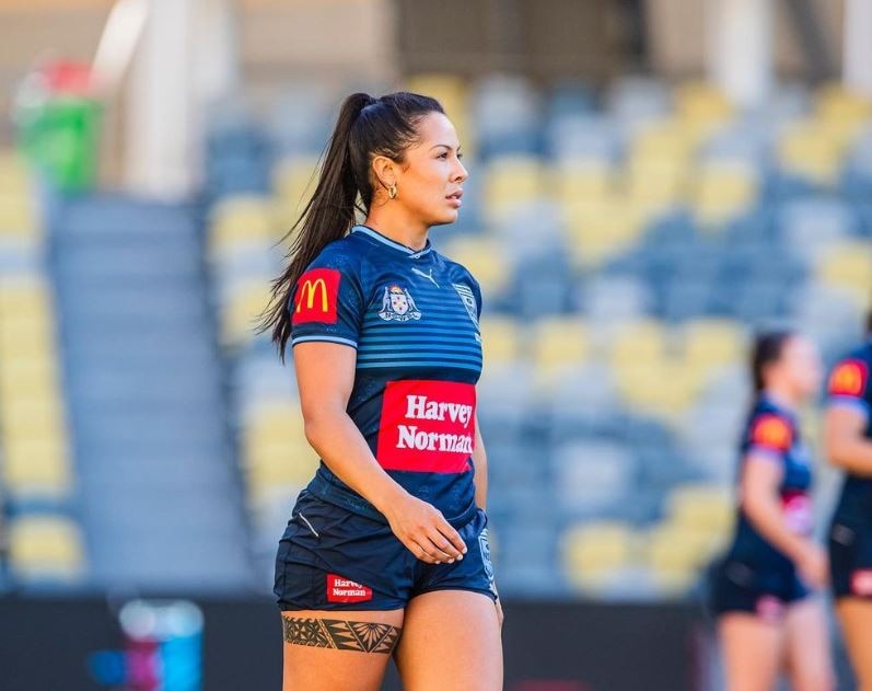 Female rugby athlete stands on stadium field in blue uniform. chairs blurred behind her. 