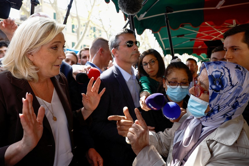 A blonde woman puts her hands up as she speaks to an older woman in a head