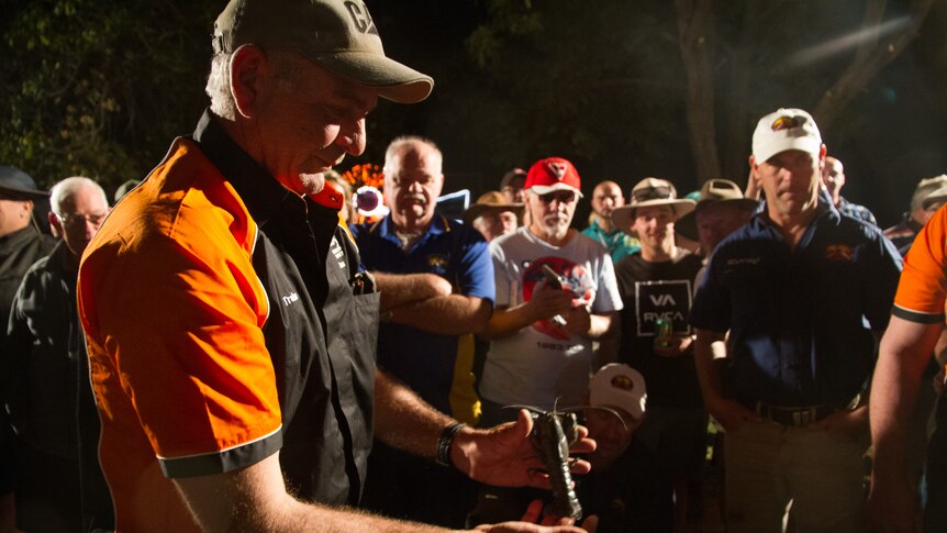 A man holds a large grey-green yabbie in his hands as a crowd of men looks on at night-time.