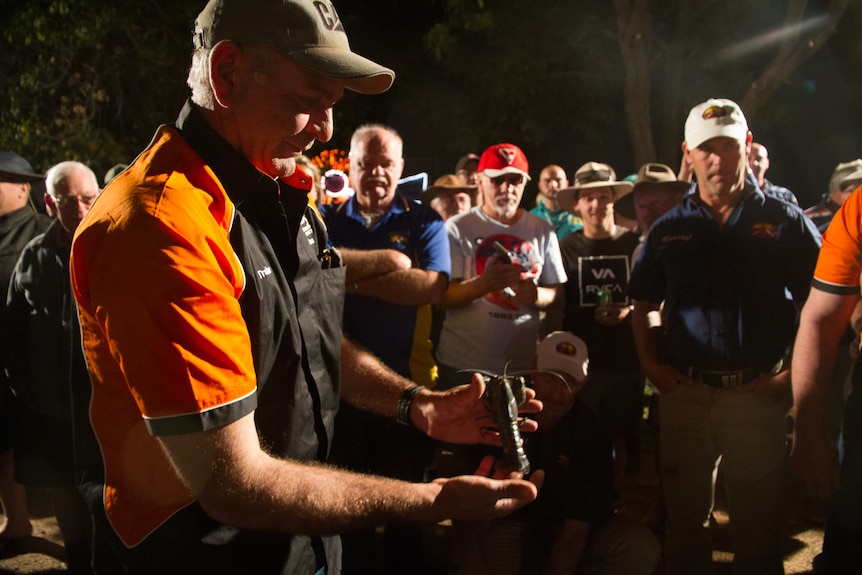 A man holds a large grey-green yabbie in his hands as a crowd of men looks on at night-time.