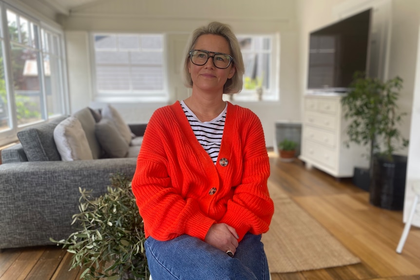 A woman with short blonde hair and glasses wearing a red cardigan sits in a living room.