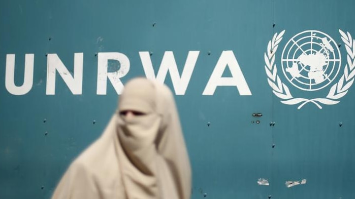 Palestinian woman in a burqa walks in front of a white UNRWA logo on a green background
