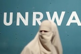 Palestinian woman in a burqa walks in front of a white UNRWA logo on a green background