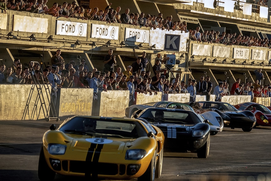 Spectators stand cheering from racing track stands at a line-up of five Ford GT40 racing cars during golden hour.