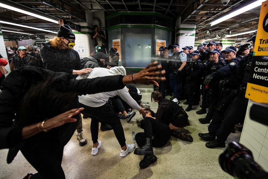 A police officer sprays capsicum spray towards protesters in a train station.