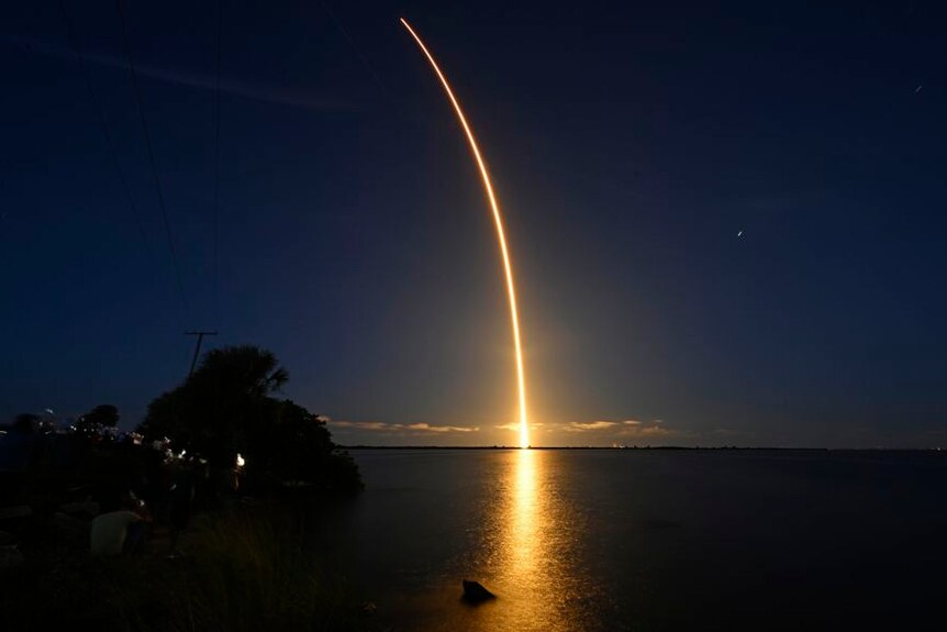 A bright orange light from a rocket take off projects across a moonlit sky at night