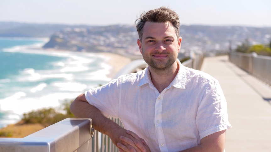 Paul Culliver, in a white shirt, leaning on the railing overlooking a white sand beach, Newcastle in the background.