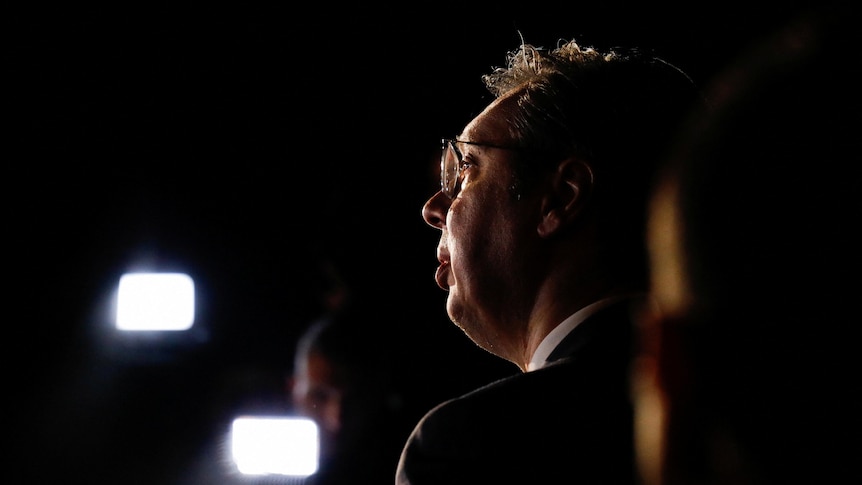 A profile shot of a middle-aged white man with glasses in a suit lit by bright lights against a dark background.