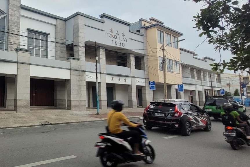 A street view, with blurred motorbikes going past, and a building with the words 'Toko Lay 1959' on it.