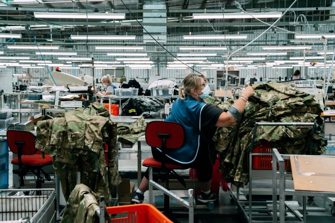 a woman reaches for a pile of army uniform khakis in a factory