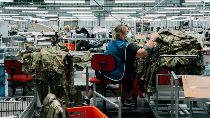 a woman reaches for a pile of army uniform khakis in a factory