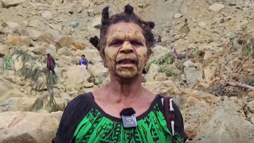 A woman in a black and lime green top, with her hair in tight knots, speaks to a camera as people behind her search in rubble
