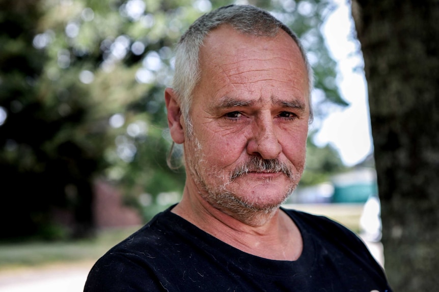 Jamie Cox with grey and black hair, moustache thin beard and black t-shirt, stands outside with trees in background