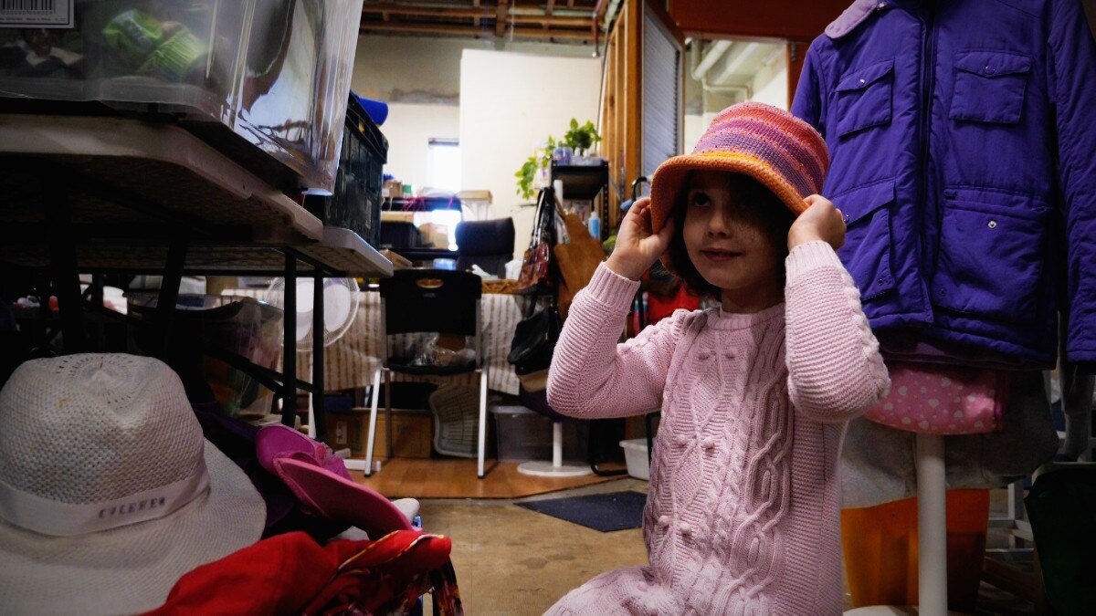 A young girl in a pink jumper trying on a colorful hat in an op shop.