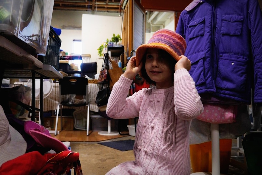A young girl in a pink jumper trying on a colorful hat in an op shop.