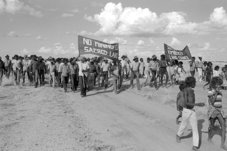 A black and white photo of Indigenous people marching across a dusty paddock, holding banners.