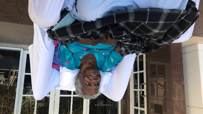 An Indigenous woman lies in a seated bed outside a court building. She smiles and looks off-camera.