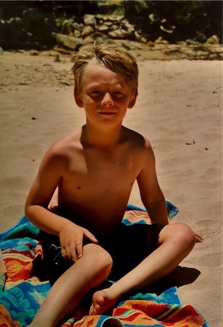 A boy in blue shorts sits on a towel on the sand.