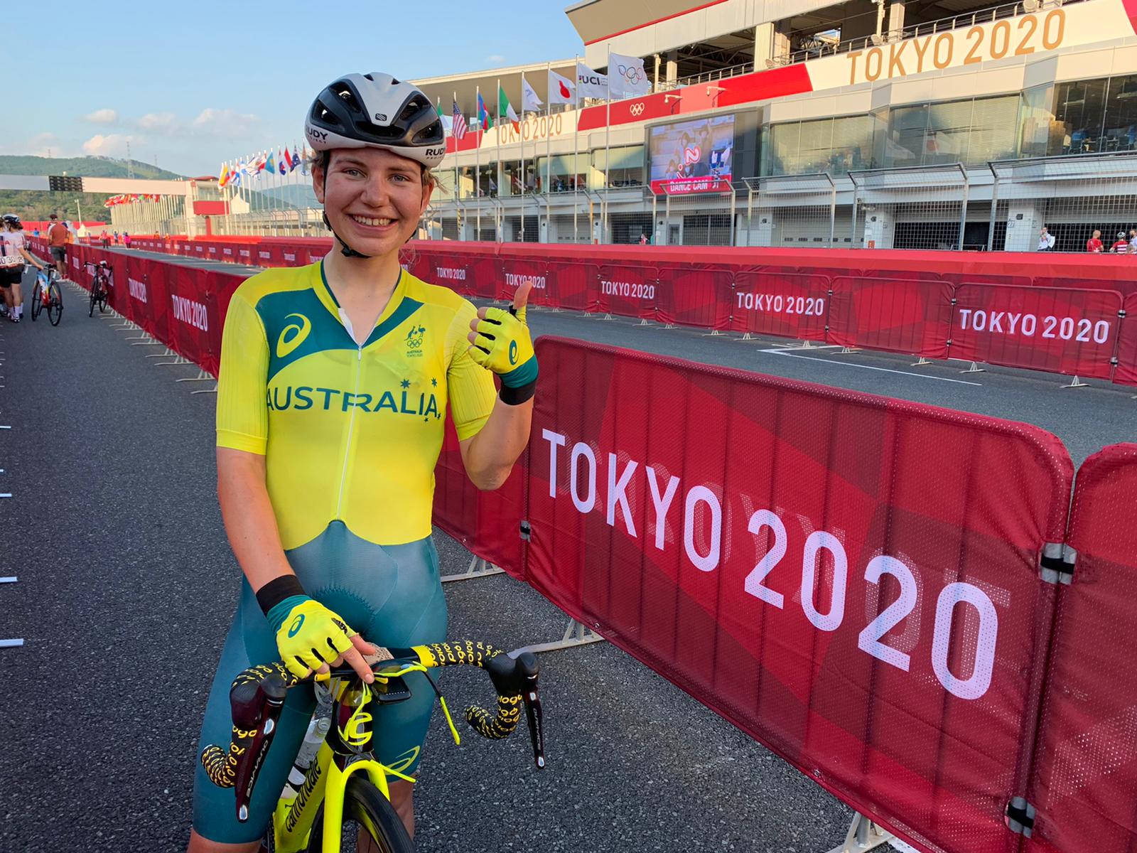 Sarah Gigante rests on her bike in front of a Tokyo Olympics sign and gives a thumbs up.