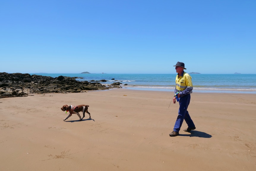 Tom Garrett and Rocky the dog walking on sand, rocks, ocean and blue sky behind.