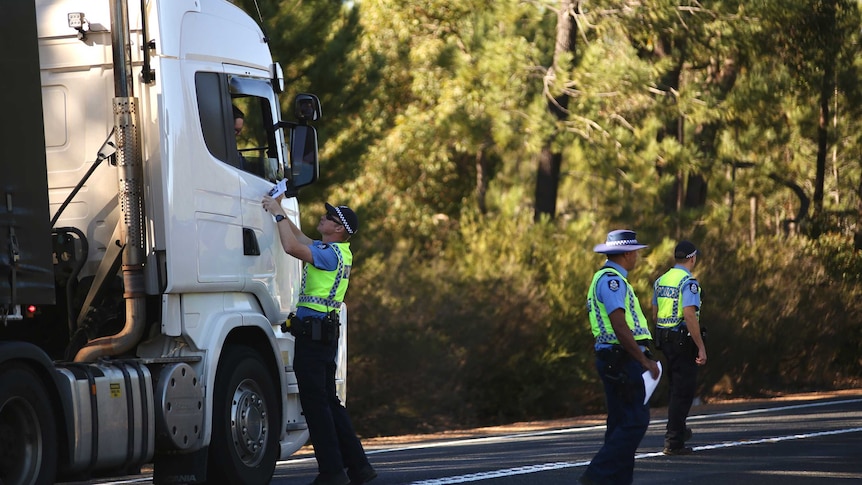 A police officer stands on a road showing a flyer to a truck driver as two other police officers stand nearby.