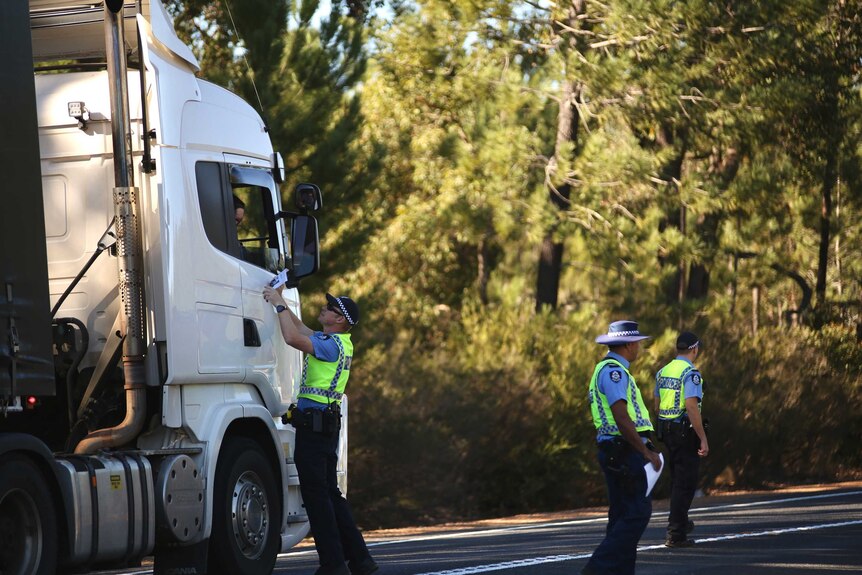 A police officer stands on a road showing a flyer to a truck driver as two other police officers stand nearby.