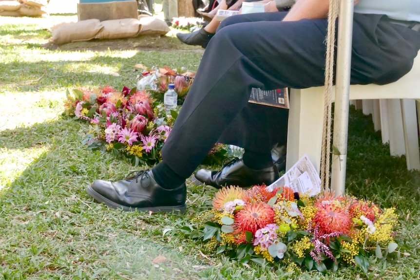 Colorful wreaths lay on the green grass next to the foot of a guest at the 80th commemorations