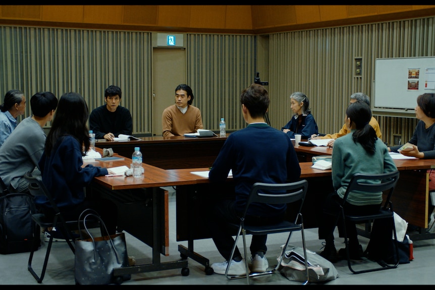Ten people sit around a square table working on a script. The room is sparse, with a whiteboard in the background.