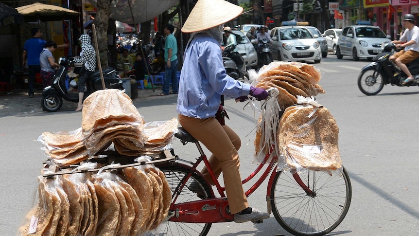A street vendor rides down a Hanoi street with large, flat rice crackers wrapped in plastic hanging from her bicycle.