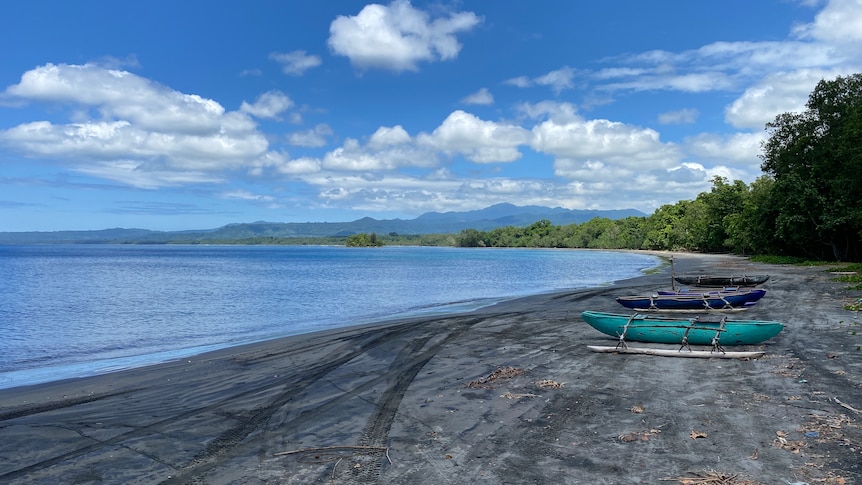 Three outrigger canoes on the shore of a black sand beach, with mountains on the horizon.