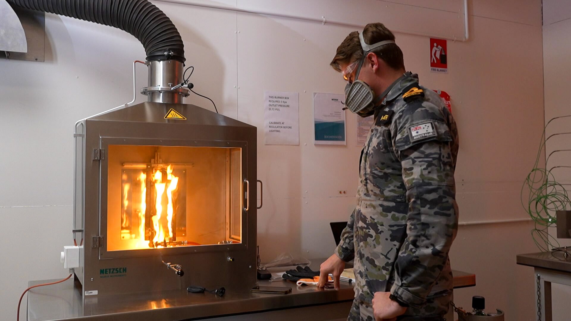 Man wearing Royal Australian Navy uniform and wearing a mask beside an oven-like device containing flames. 