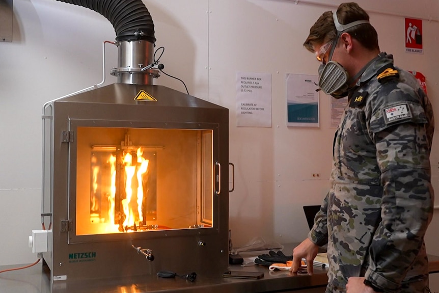 Man wearing Royal Australian Navy uniform and wearing a mask beside an oven-like device containing flames. 