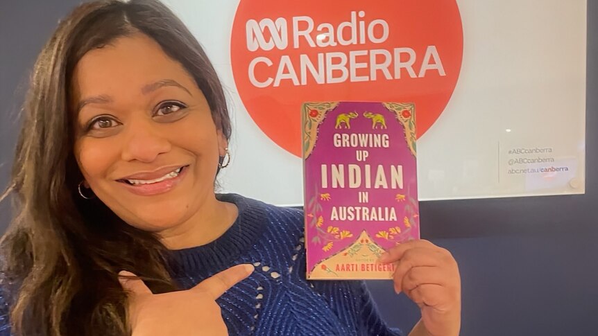 A woman in a blue top holds up a copy of her book, Growing up Indian in Australia