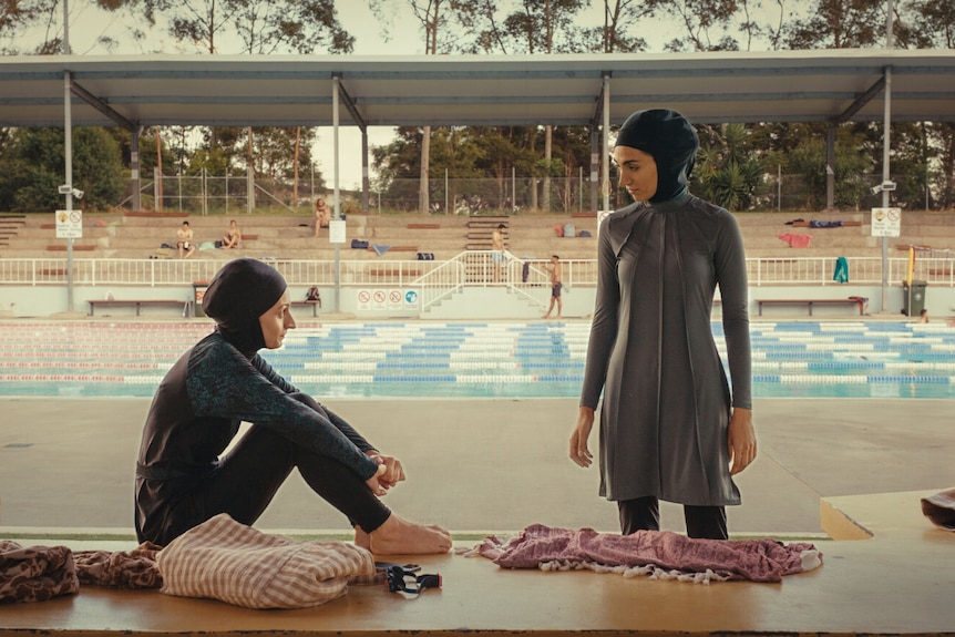 Two Muslim women sit in shade at public swimming pool wearing Lycra head scarves, tights and swimming tunics.