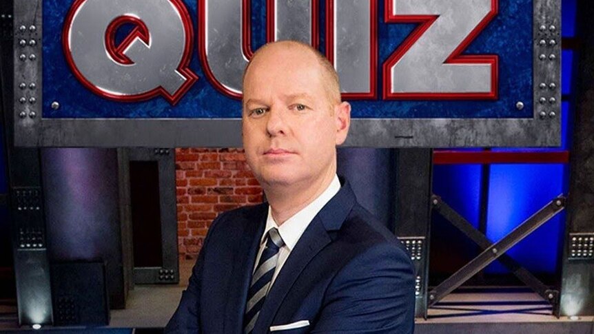 Tom Gleeson, a bald white man, wears a navy suit and stands on a TV set with a large sign reading "Hard Quiz".