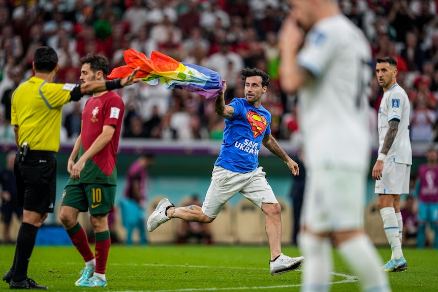 A man running across a soccer field with a rainbow flag with multiple players standing around