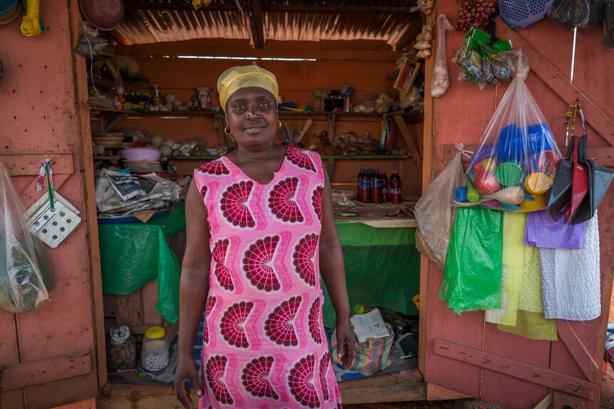 A woman wearing a bright pink and red patterned dress stands in front of a Ghanian market stall.