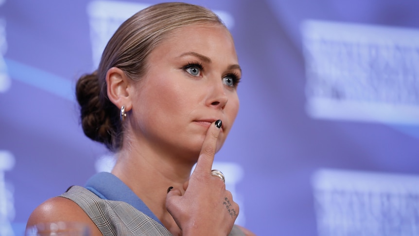 A woman with blonde hair tied back in a low bun presses a finger to her lips, nail painted black.