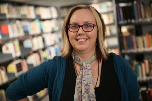 Smiling young Caucasian woman with glasses, a bob cut, wearing patterned scarf and blue cardigan in a room full of books.