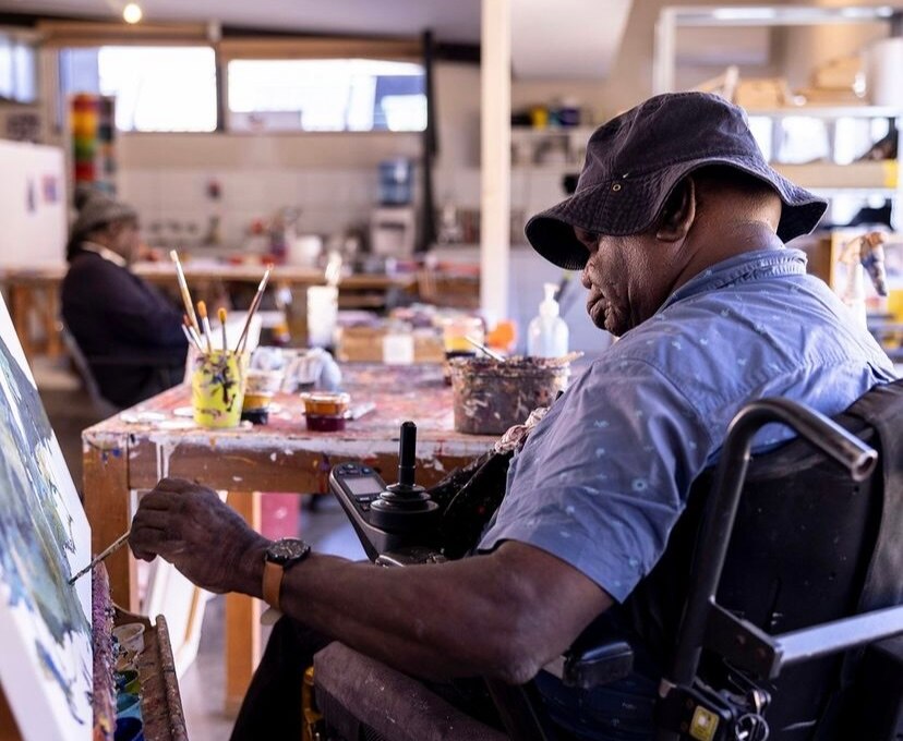 A man in a floppy hat using a wheelchair paints something onto a canvas