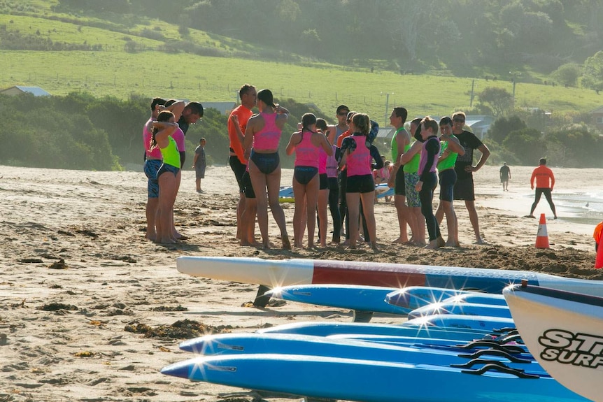 A trainer talks to a group of young surf lifesavers on the beach, paddle boards lined up in foreground