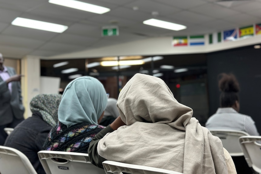 Two women wearing hijabs listen to a speaker in a meeting.
