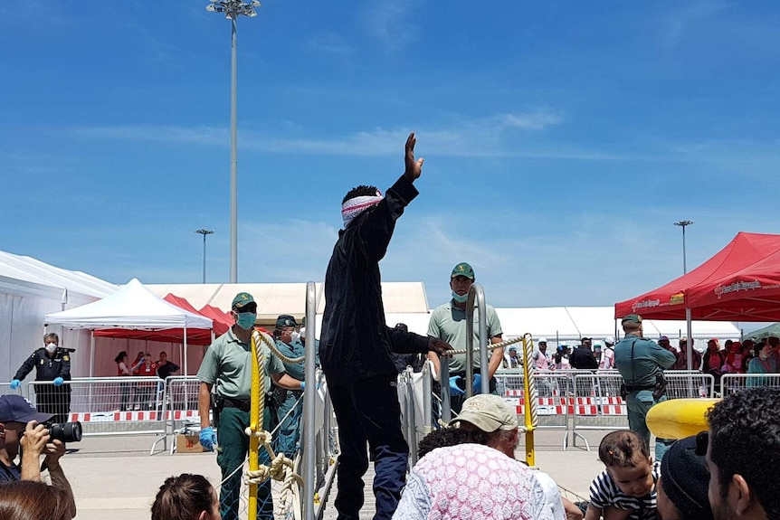 Man waves as he exits the boat, greeted by officials wearing face masks